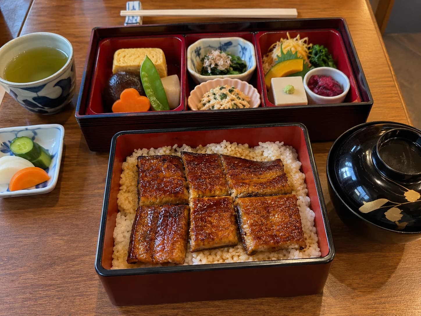 AI-generated sample guest photo: a lacquered eel rice box and seasonal bento, photographed from a seated angle